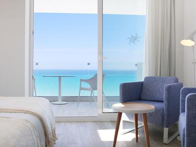 Helles Hotelzimmer mit Balkon, blauer Stuhl, Bett und Blick aufs Meer.