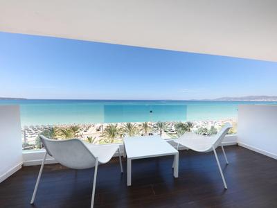 Balkon mit zwei weißen Stühlen und Tisch mit Blick auf das Meer und Palmen.