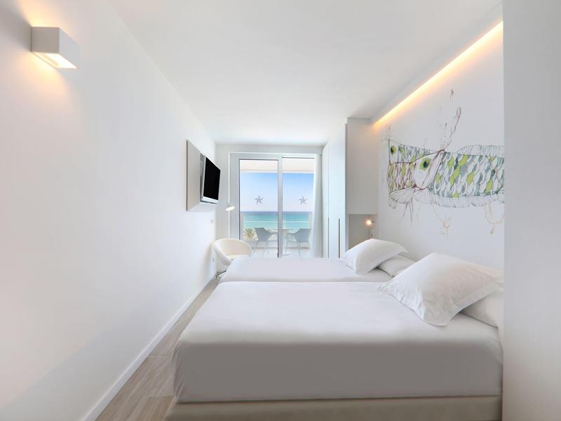 Habitación de hotel luminosa con dos camas, TV en la pared y vista al mar desde ventanas grandes.