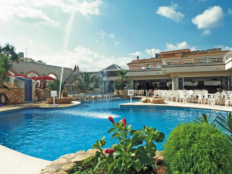 Hotelový bazén s sluneční terasou, posezením a bujnou zelení pod modrou oblohou.