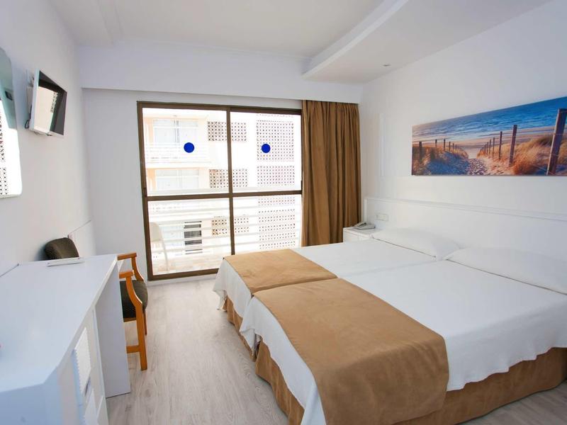 Světle zařízený hotelový pokoj se dvěma postelemi a velkým oknem s výhledem na sousední budovy.