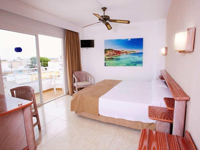 Světle zařízený hotelový pokoj s manželskou postelí, balkonem a výhledem na moře.