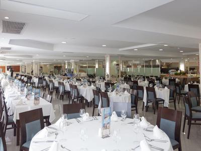 Grande salle à manger lumineuse avec tables dressées en blanc et chaises marron dans un hôtel.