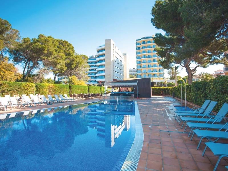 Hôtel avec grande piscine extérieure, chaises longues et arbres sous un ciel clair.