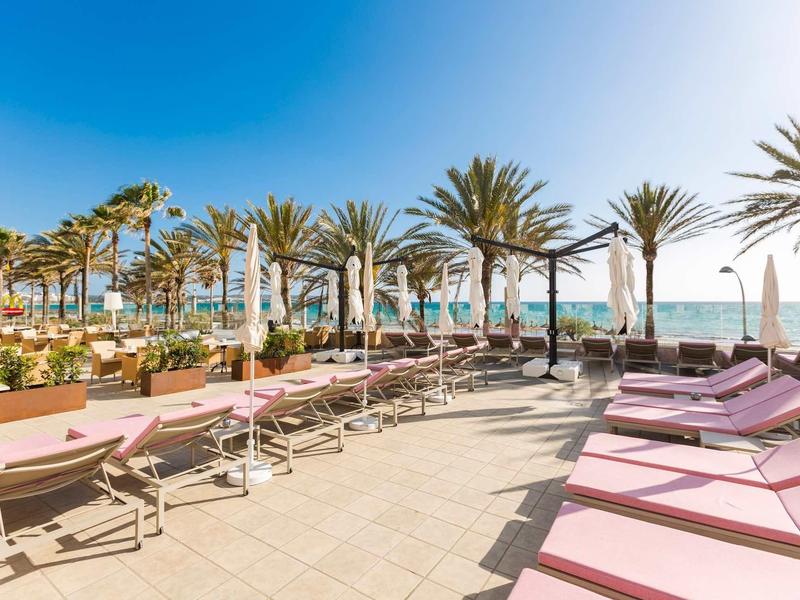 Słoneczny taras z leżakami, palmami i widokiem na morze w hotelu nad plażą.