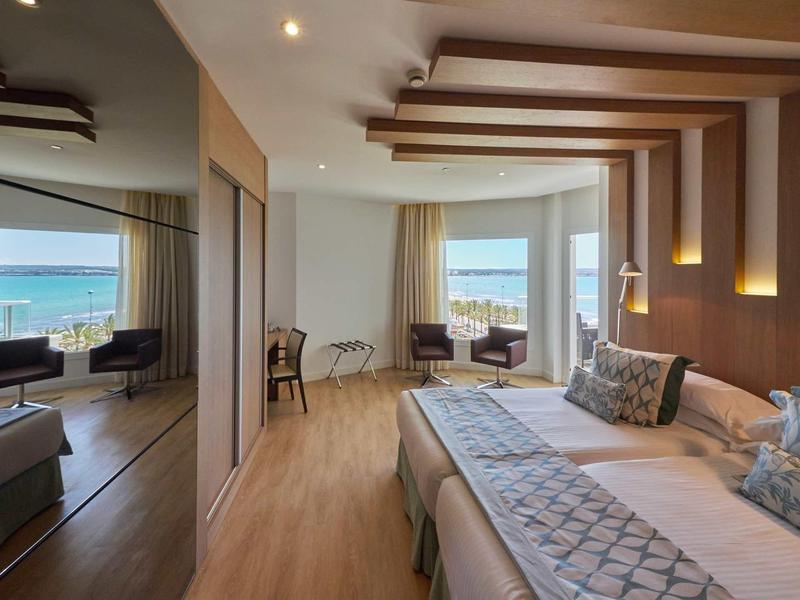 Nowoczesny pokój hotelowy z dużym łóżkiem, balkonem i widokiem na morze w świetle dziennym.