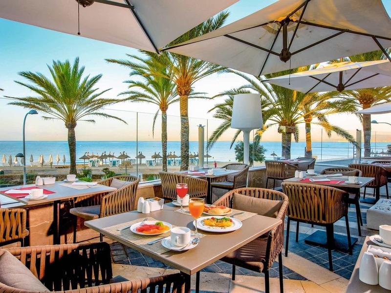 Taras z nakrytymi stołami, parasolami, palmami i widokiem na morze o zachodzie słońca.