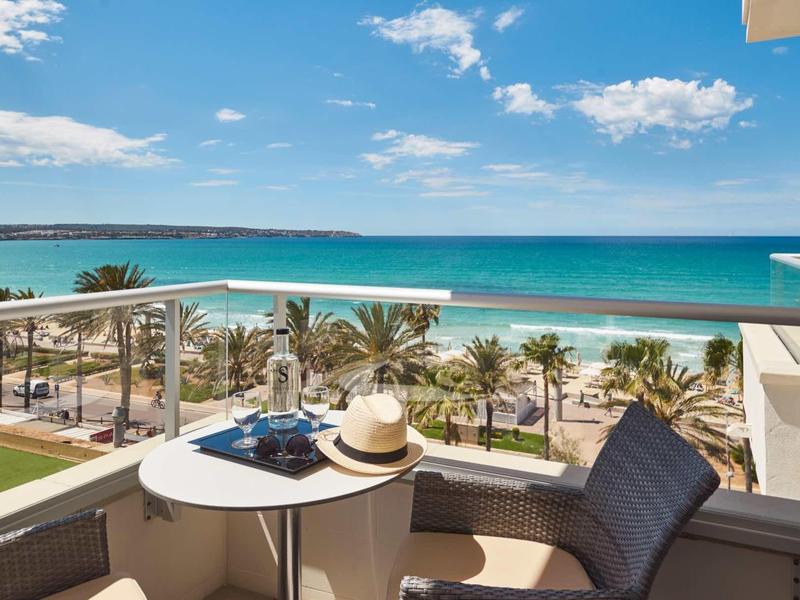 Balkon z krzesłem, stołem i widokiem na plażę, palmy i niebieskie morze pod niebieskim niebem.