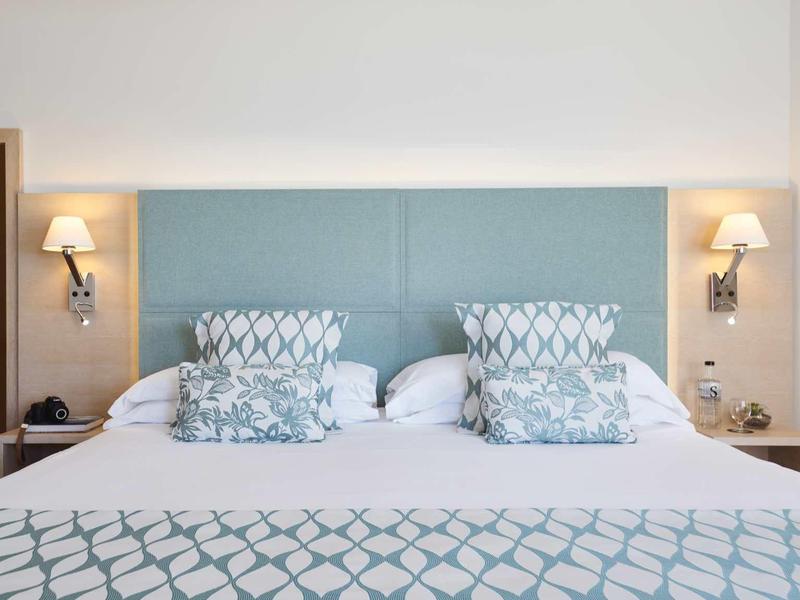 Nowoczesny pokój hotelowy z podwójnym łóżkiem, tapicerowanym zagłówkiem i symetrycznymi lampami ściennymi.