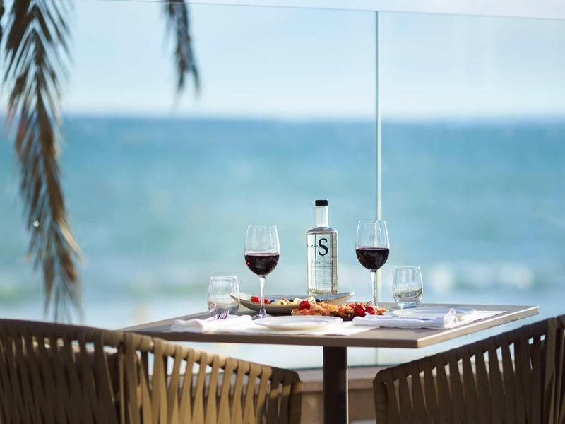 Stół zastawiony winem i wodą z widokiem na morze i palmy w tle.