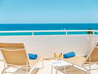 Zwei beige Sonnenliegen mit blauen Handtüchern auf weißer Terrasse mit Meerblick und klarem Himmel.