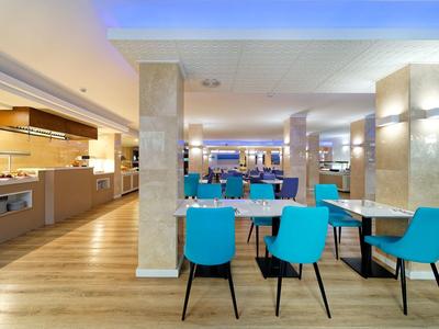 Moderner Speisesaal mit blauen Stühlen, rechteckigen Tischen und hellem Holzfußboden.