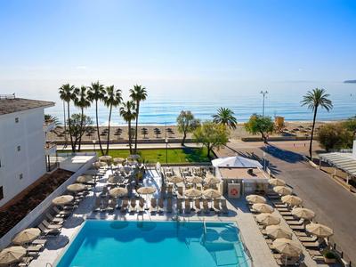 Pool mit Sonnenliegen am Strand, Palmen und ruhigem Meer im Hintergrund an klarem Tag.