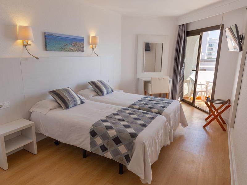 Bright hotel room with two single beds, balcony, TV, and wooden flooring.