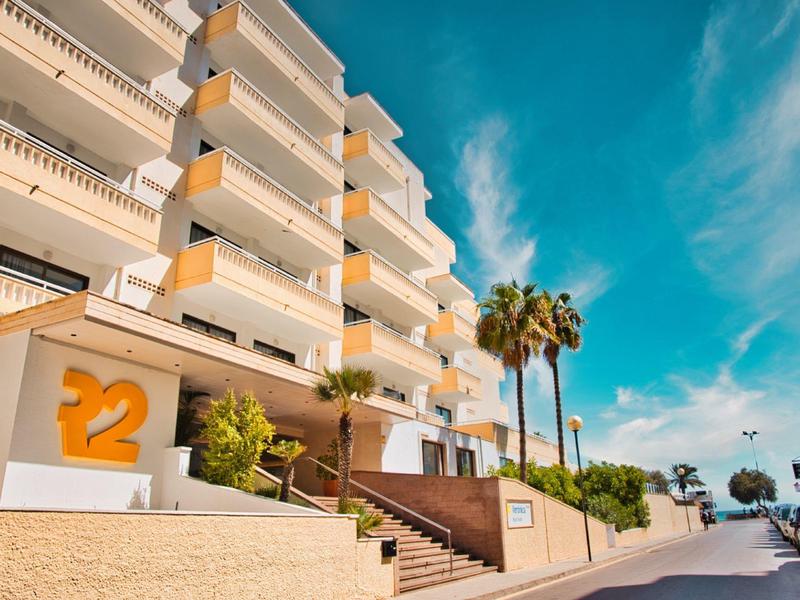 Modern hotel exterior with balconies, palm trees, and clear blue sky on a sunny day.