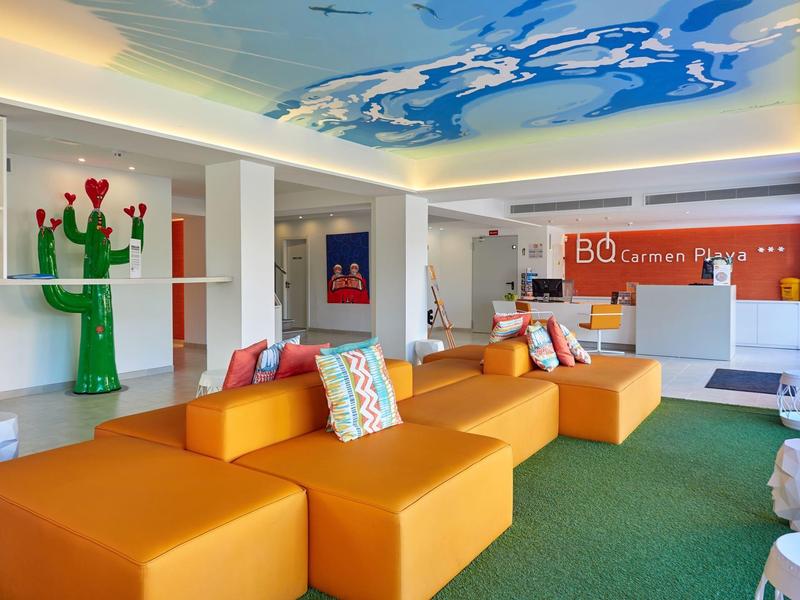 Área moderna de lobby de hotel con sofás naranjas, cojines coloridos y alfombra verde.