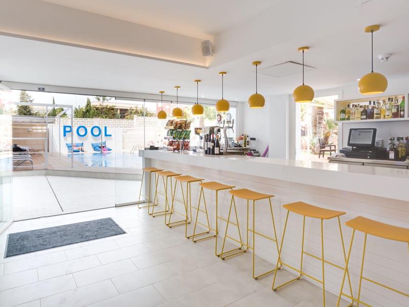 Moderna área de bar de hotel con sillas blancas y amarillas y vista a la piscina al fondo.