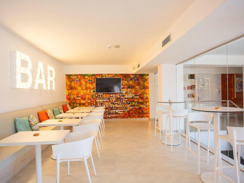 Área de bar luminosa con mesas y sillas blancas y banco colorido frente a pared de piedra con televisor.