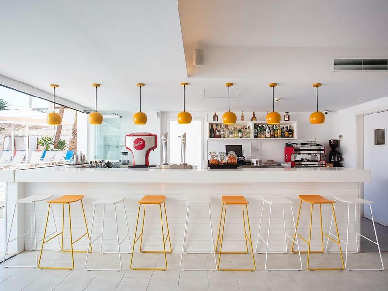 Bar moderna con barras blancas, taburetes amarillos y lámparas colgantes naranjas en un espacio luminoso.