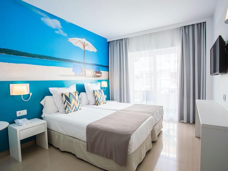 Habitación de hotel moderna con dos camas individuales, mural azul de playa y gran ventana con cortinas.