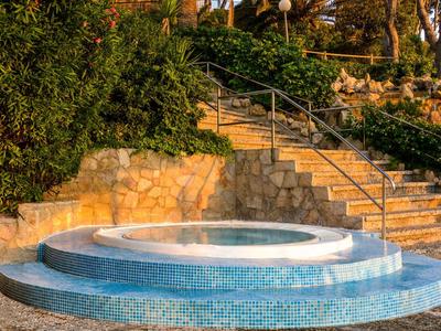 Okrągła fontanna z niebieską mozaiką, otoczona kamiennymi schodami i zielenią.