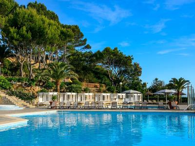 Jasnoniebieski basen z leżakami i parasolami, otoczony bujną roślinnością pod błękitnym niebem.