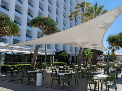 Zewnętrzny obszar hotelu z żaglem przeciwsłonecznym oraz wieloma stołami i krzesłami.