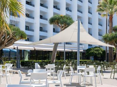 Nowoczesny teren na zewnątrz z białymi krzesłami, stolikami i dużym daszkiem przeciwsłonecznym przed hotelem.