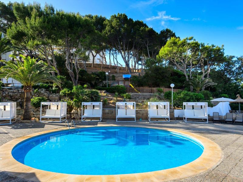 Okrągły basen z białymi leżakami i drzewami w tle pod niebieskim niebem.