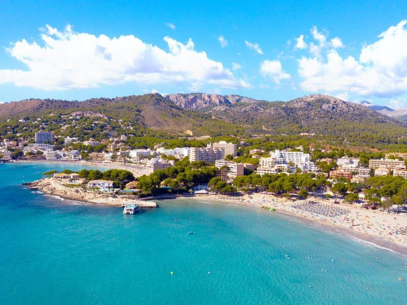 Piaszczysta plaża z niebieską wodą i górami w tle pod częściowo pochmurnym niebem.