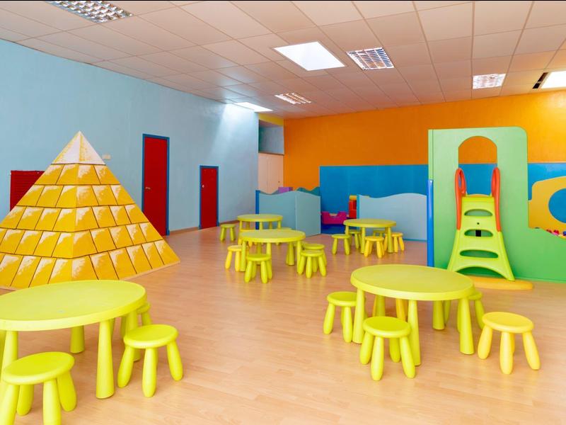 Kolorowa sala zabaw z żółtymi okrągłymi stolikami i wewnętrznym placem zabaw dla dzieci.
