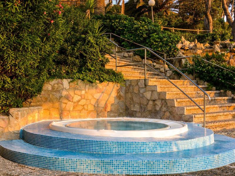 Okrągła fontanna z niebieską mozaiką, otoczona kamiennymi schodami i zielenią.