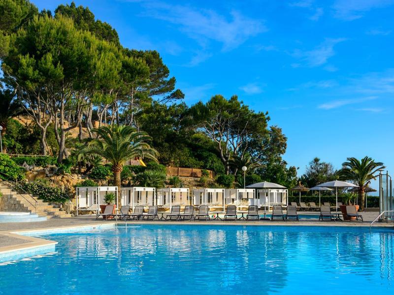 Jasnoniebieski basen z leżakami i parasolami, otoczony bujną roślinnością pod błękitnym niebem.