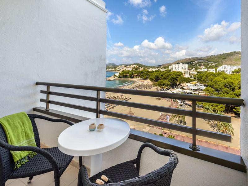Balkon hotelowy z białym owalnym stołem, dwoma czarnymi krzesłami i widokiem na morze i wzgórza.