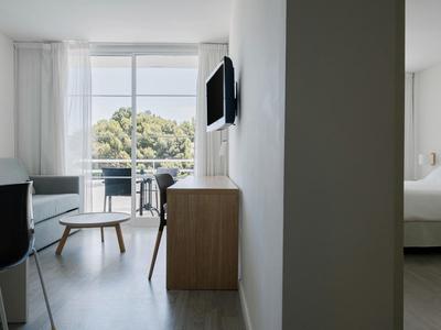 Nowoczesny pokój hotelowy z balkonem, sofą, stołem i widokiem na zielone drzewa.