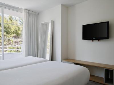 Jasny pokój hotelowy z łóżkiem podwójnym, dużym lustrem, telewizorem na ścianie i widokiem na zielone drzewa.