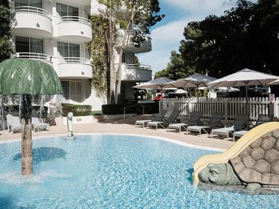 Basen hotelowy z zjeżdżalnią wodną, fontanną w kształcie grzyba i leżakami pod parasolami.