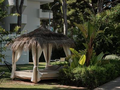 Overdekte dagbed met gordijnen staat in de tuin naast struiken en palmbomen.