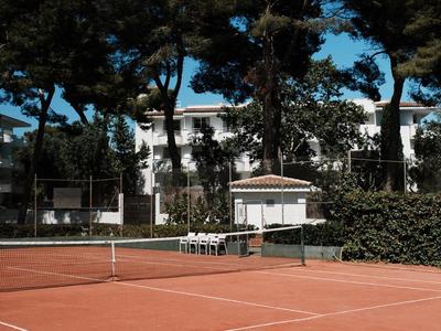 Tennisbaan met rode gravel voor bomen onder een blauwe lucht.