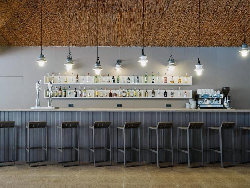 Nowoczesny bar hotelowy z szarymi stołkami barowymi i półkami na butelki pod drewnianym sufitem.