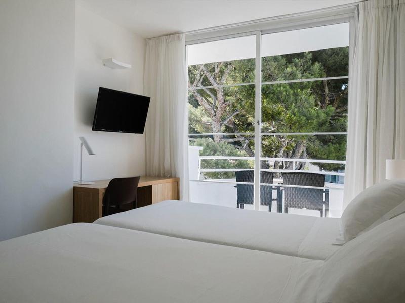 Jasny pokój hotelowy z dwoma pojedynczymi łóżkami i balkonem z widokiem na zielone otoczenie.