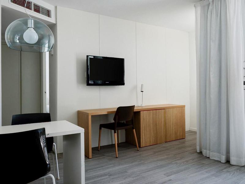 Jasny pokój hotelowy z biurkiem, krzesłem, telewizorem na ścianie i zasłonami przy oknie.