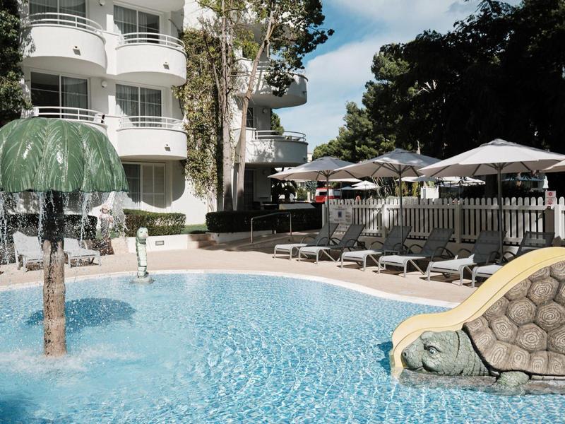 Basen hotelowy z zjeżdżalnią wodną, fontanną w kształcie grzyba i leżakami pod parasolami.