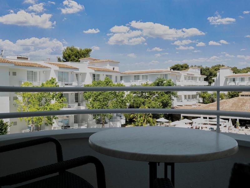 Widok z balkonu z stołem i krzesłami na budynek hotelu i drzewa pod niebieskim niebem.