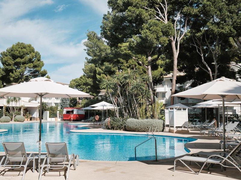 Basen z leżakami i parasolami otoczony drzewami pod niebieskim niebem.