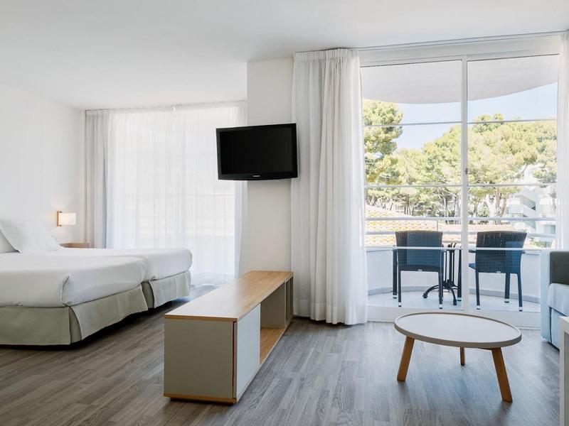 Jasny pokój hotelowy z podwójnym łóżkiem, telewizorem, częścią wypoczynkową i balkonem z widokiem na drzewa.