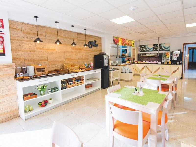 Hell eingerichteter Frühstücksraum mit Holzakzenten, Tischen und Stühlen, Kühlschrank und Buffetbereich.