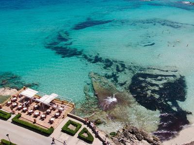 Klares türkisfarbenes Meer mit hellem Sandstrand und Terrasse eines Hotels am Ufer.