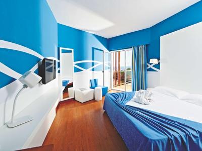 Bright hotel room with blue and white decor, double bed, seating area, and balcony with ocean view.