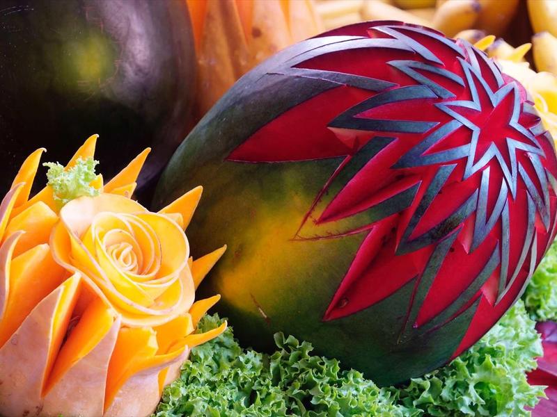 Intricately carved watermelon and yellow fruit with leafy greens background.
